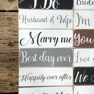 May include: Wooden rectangular signs with white script text on various colored backgrounds. Phrases include "I Do", "Husband & Wife", "Marry me", "Best day ever", "Happily ever after", and "Mr. & Mrs.". The signs are arranged on a wooden surface.