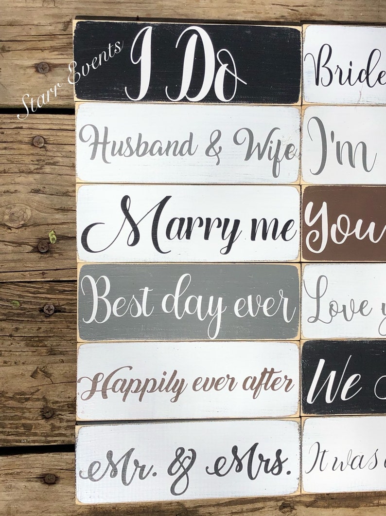 May include: Wooden rectangular signs with white script text on various colored backgrounds. Phrases include "I Do", "Husband & Wife", "Marry me", "Best day ever", "Happily ever after", and "Mr. & Mrs.". The signs are arranged on a wooden surface.