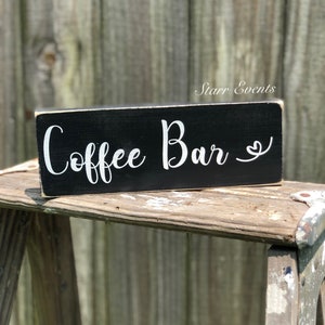 May include: A rectangular black wooden sign with the words "Coffee Bar" in white script. A small heart is next to the word "Bar". The sign is displayed on a rustic wooden surface.