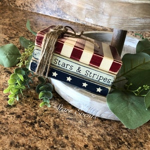 Puede incluir: Una pila de bloques de madera decorativos con tem&aacute;tica patri&oacute;tica. Los bloques est&aacute;n pintados con rayas rojas y blancas, azul con estrellas blancas y las palabras "Stars & Stripes". Atados con hilo, descansan sobre una bandeja escalonada con vegetaci&oacute;n.