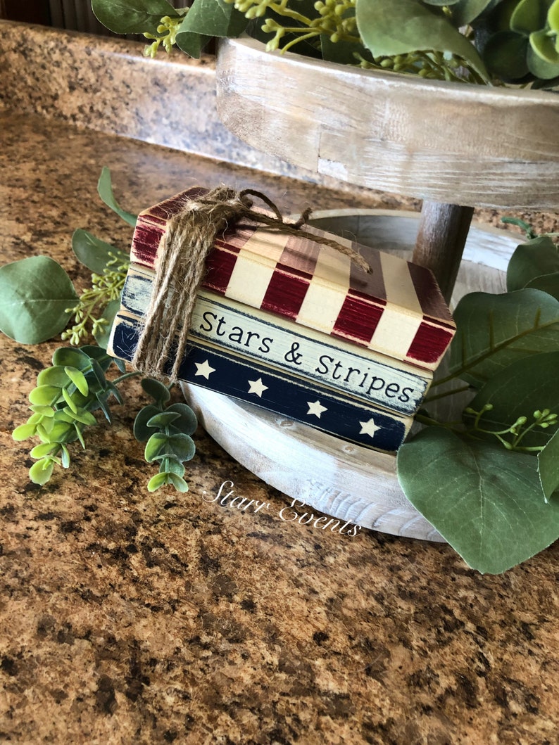 Puede incluir: Una pila de bloques de madera decorativos con tem&aacute;tica patri&oacute;tica. Los bloques est&aacute;n pintados con rayas rojas y blancas, azul con estrellas blancas y las palabras "Stars & Stripes". Atados con hilo, descansan sobre una bandeja escalonada con vegetaci&oacute;n.
