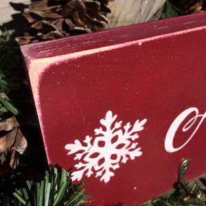 Pode incluir: Close-up de uma placa de madeira vermelha com um design de floco de neve branco. A placa tem um acabamento desgastado e um pequeno fecho de metal. A placa &eacute; cercada por agulhas de pinheiro e pinhas, sugerindo um tema de feriado.