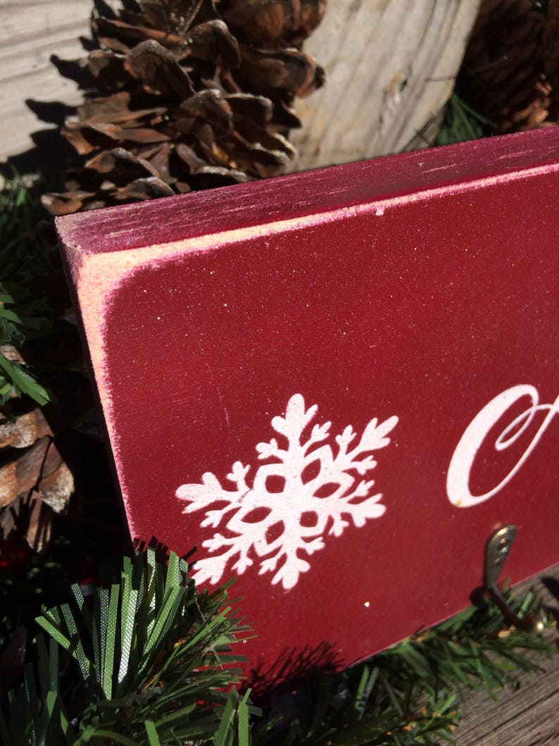 Pode incluir: Close-up de uma placa de madeira vermelha com um design de floco de neve branco. A placa tem um acabamento desgastado e um pequeno fecho de metal. A placa &eacute; cercada por agulhas de pinheiro e pinhas, sugerindo um tema de feriado.