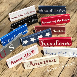 May include: A selection of rectangular wooden signs painted in red, white, and blue, each bearing a patriotic phrase. Phrases include "Land that I love," "Home of the free," and "Freedom." The signs have a distressed, rustic finish.