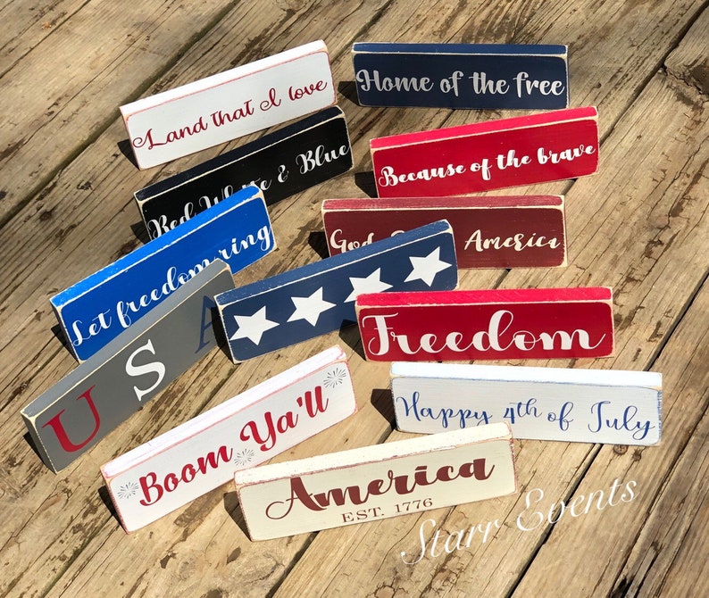 May include: A selection of rectangular wooden signs painted in red, white, and blue, each bearing a patriotic phrase. Phrases include "Land that I love," "Home of the free," and "Freedom." The signs have a distressed, rustic finish.