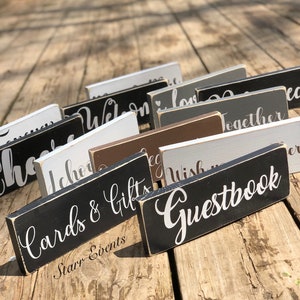 May include: A selection of rectangular wooden signs with white script lettering. The signs are painted in various colours, including black, white, grey, and brown. The signs read "Cards & Gifts", "Guestbook", and other event-related phrases.