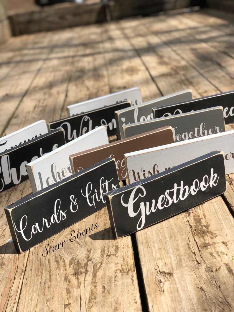 May include: A selection of rectangular wooden signs with white script lettering. The signs are painted in various colours, including black, white, grey, and brown. The signs read "Cards & Gifts", "Guestbook", and other event-related phrases.