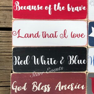 May include: A stack of wooden blocks painted in red, white, and blue, each bearing a white script message. Phrases include "Home of the free" and "Let freedom ring." The rustic style evokes a sense of patriotic sentiment.