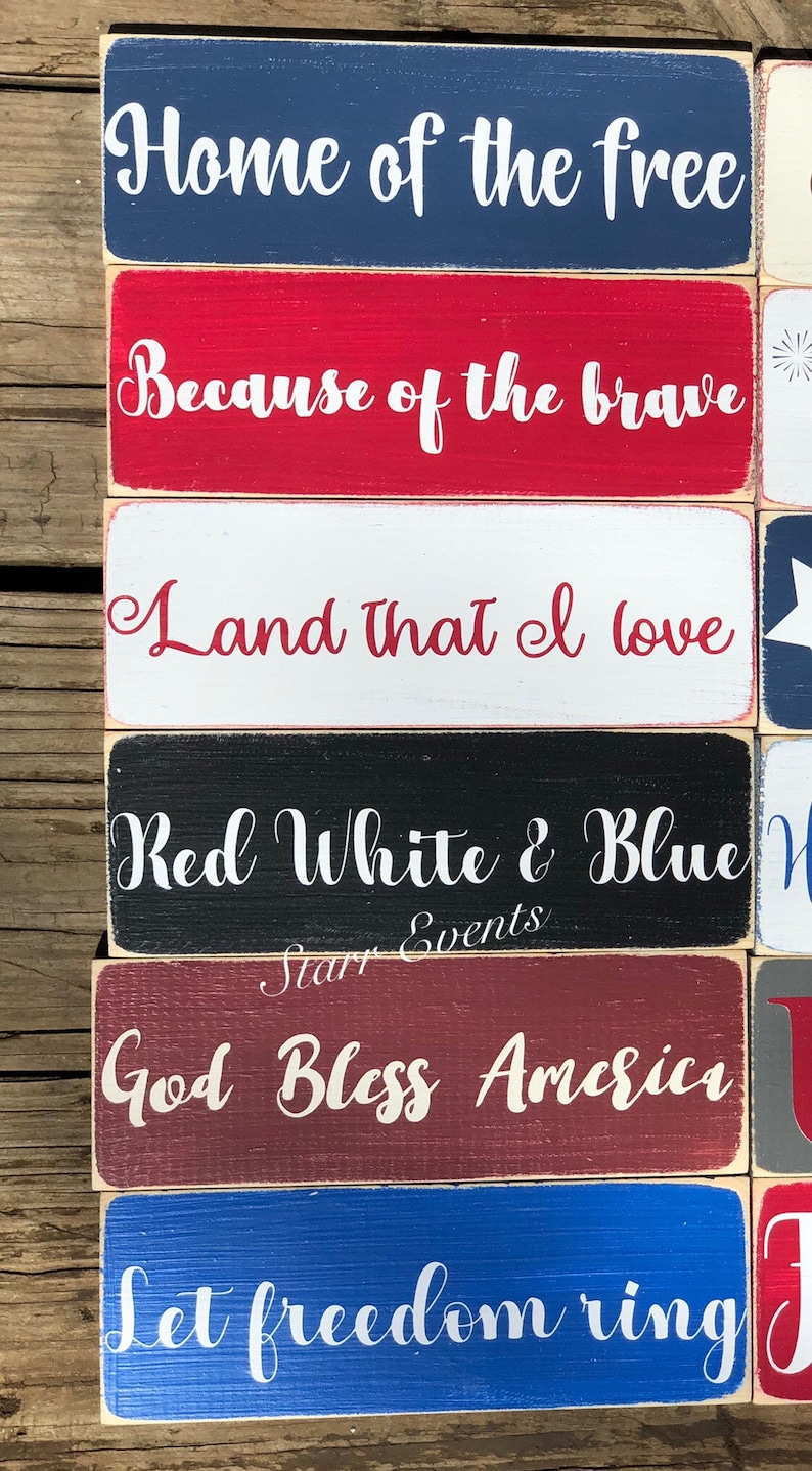 May include: A stack of wooden blocks painted in red, white, and blue, each bearing a white script message. Phrases include "Home of the free" and "Let freedom ring." The rustic style evokes a sense of patriotic sentiment.