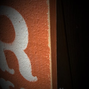 May include: Close-up of a wooden sign with a textured orange surface and white lettering. The edge of the sign is visible, showing a lighter colour. The background is dark.