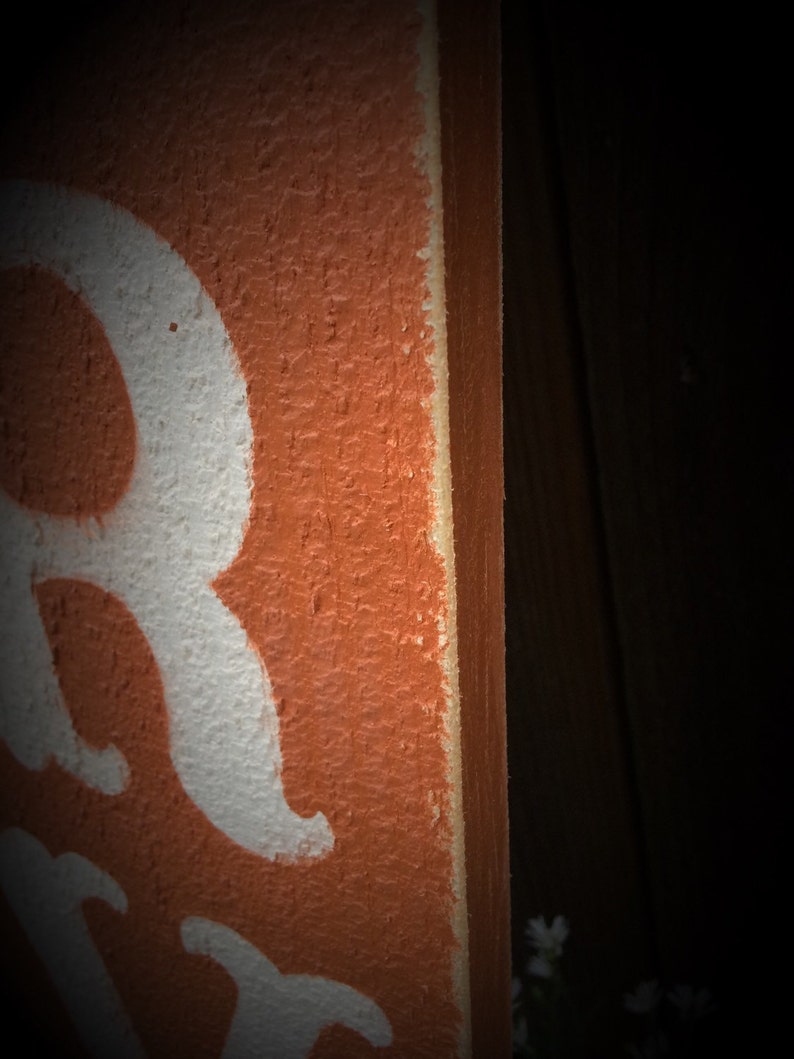 May include: Close-up of a wooden sign with a textured orange surface and white lettering. The edge of the sign is visible, showing a lighter colour. The background is dark.