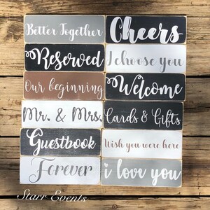 May include: A selection of rectangular wooden signs with white lettering on various coloured backgrounds. Phrases include "Better Together," "Cheers," "Reserved," "I Choose You," "Our Beginning," and "Welcome." The signs are arranged on a wooden surface.