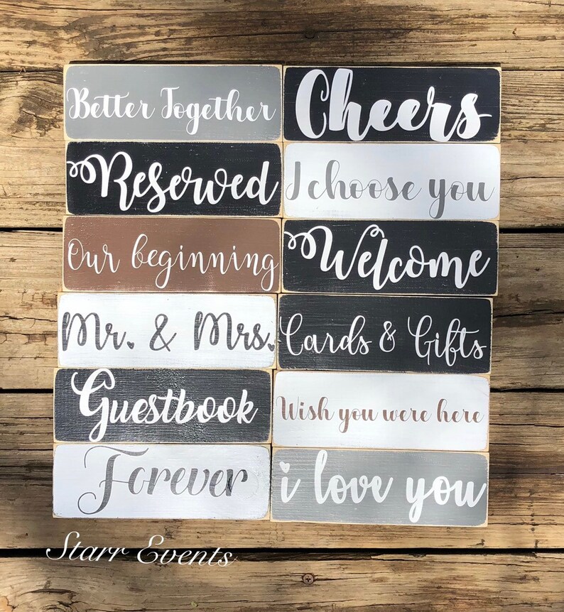 May include: A selection of rectangular wooden signs with white lettering on various coloured backgrounds. Phrases include "Better Together," "Cheers," "Reserved," "I Choose You," "Our Beginning," and "Welcome." The signs are arranged on a wooden surface.