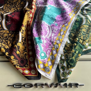 May include: Three square scarves featuring intricate baroque-style scrollwork, engine piston motifs, and geometric diamond borders. The scarves are colored in black and gold, white with purple and teal, and dark green with gold, resting on a Corvair emblem.