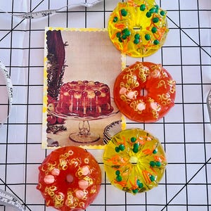 Puede incluir: Cuatro adornos circulares de resina translúcida con forma de moldes de anillo, dos amarillos con inclusiones verdes y naranjas y dos naranjas con inclusiones rosas y doradas, junto a una postal antigua y una cinta métrica blanca.