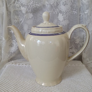 May include: A cream-colored ceramic coffee pot with a blue trim around the lid and spout. The pot has a classic design with a rounded body and a tall, slender spout.
