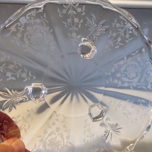 May include: A clear glass pedestal bowl with a scalloped edge and etched floral design. The bowl has three feet.