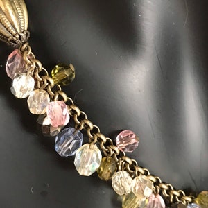 Puede incluir: Un collar de cadena dorada con un colgante grande y adornado y un racimo de cuentas de vidrio de colores. Las cuentas son en tonos de rosa, azul, verde y transparente.