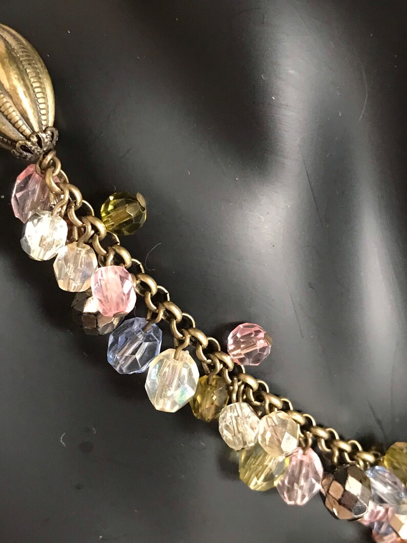 Puede incluir: Un collar de cadena dorada con un colgante grande y adornado y un racimo de cuentas de vidrio de colores. Las cuentas son en tonos de rosa, azul, verde y transparente.