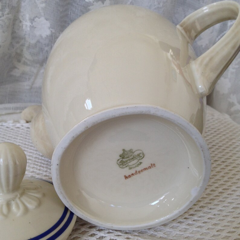 Pu&ograve; includere: Una teiera in ceramica bianca con un coperchio e un manico. La teiera ha un logo verde e oro sul fondo che recita "Bavaria" e "handgemalt".