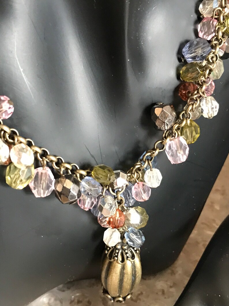 Puede incluir: Un collar de cadena dorada con una variedad de cuentas de vidrio de colores y un colgante decorativo de oro. Las cuentas son en tonos de rosa, azul, verde y marr&oacute;n.