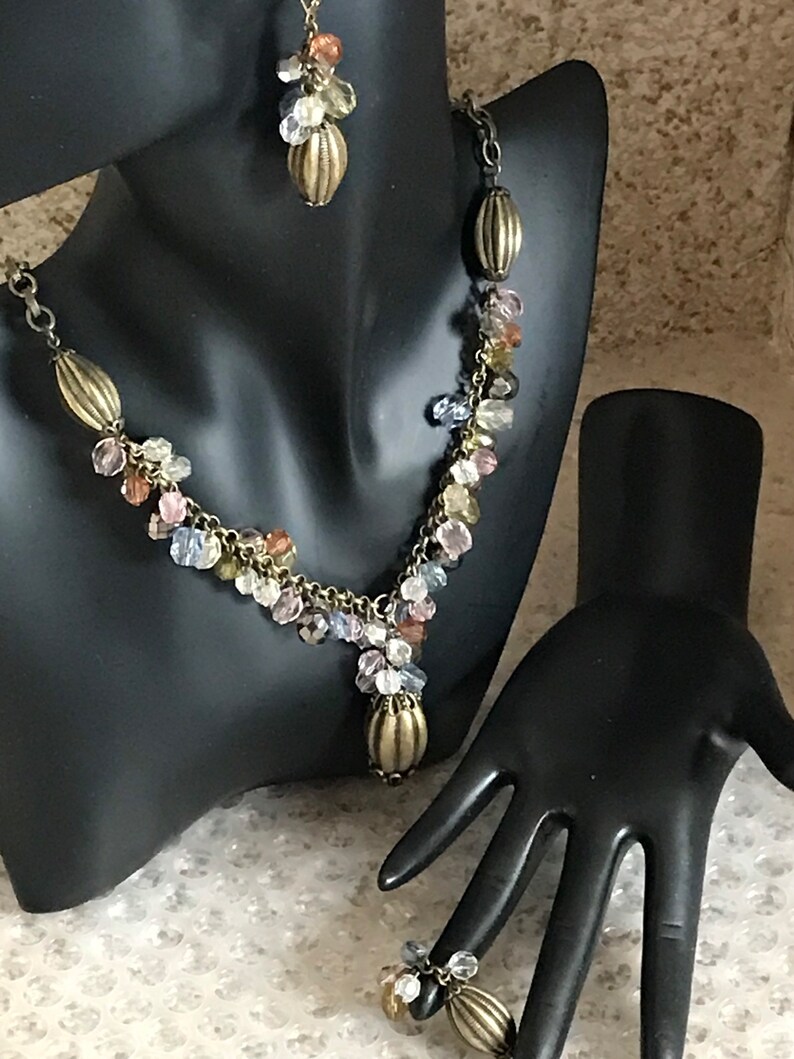 Puede incluir: Un collar de m&uacute;ltiples hilos y pendientes a juego con una cadena de bronce y cuentas de cristal de colores. El collar tiene un colgante en forma de l&aacute;grima con una tapa de bronce. Tambi&eacute;n se muestra un anillo a juego con una tapa de bronce y cuentas de cristal.