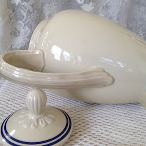 Pu&ograve; includere: Un'anfora in ceramica bianca con un coperchio a strisce blu e bianche. L'anfora ha un manico e un pomo decorativo sul coperchio.