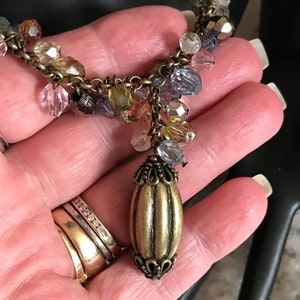 Puede incluir: Un collar dorado con una cadena de cuentas y un gran colgante adornado. Las cuentas son multicolores e incluyen tonos de rosa, azul, verde y marr&oacute;n.