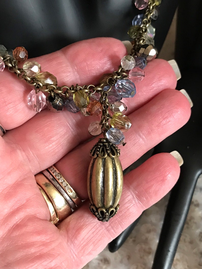 Puede incluir: Un collar dorado con una cadena de cuentas y un gran colgante adornado. Las cuentas son multicolores e incluyen tonos de rosa, azul, verde y marr&oacute;n.