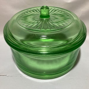 May include: A round, light green glass bowl with a matching lid. The lid features a central knob and a radiating sunburst pattern of embossed lines. The bowl has smooth, rounded sides and a translucent finish, showcasing the vibrant green color.