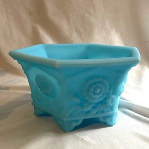 May include: A light blue, square-shaped planter with a floral design on the sides. The planter has a textured surface and sits on four small feet. The top edge has a slightly raised rim.
