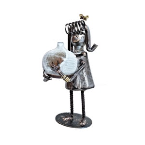 May include: A metal figurine of a person holding a white ceramic vase. The figurine is made of recycled metal and has a rustic, industrial look. The vase has a textured surface and is a unique piece of home decor.