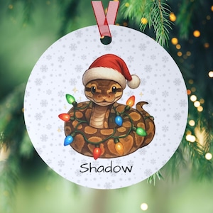 May include: A round Christmas ornament featuring a snake wearing a Santa hat and wrapped in colorful Christmas lights. The ornament has a snowflake pattern and the name "Shadow" printed on it. A red ribbon is attached for hanging.
