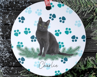 Könnte beinhalten: Runder weißer Weihnachtsbaumschmuck mit einer schwarzen Katze, die auf einem grünen Tannenzweig sitzt. Der Baumschmuck hat ein blaues Pfotenabdruckmuster und der Name "Charlie" ist unten geschrieben.