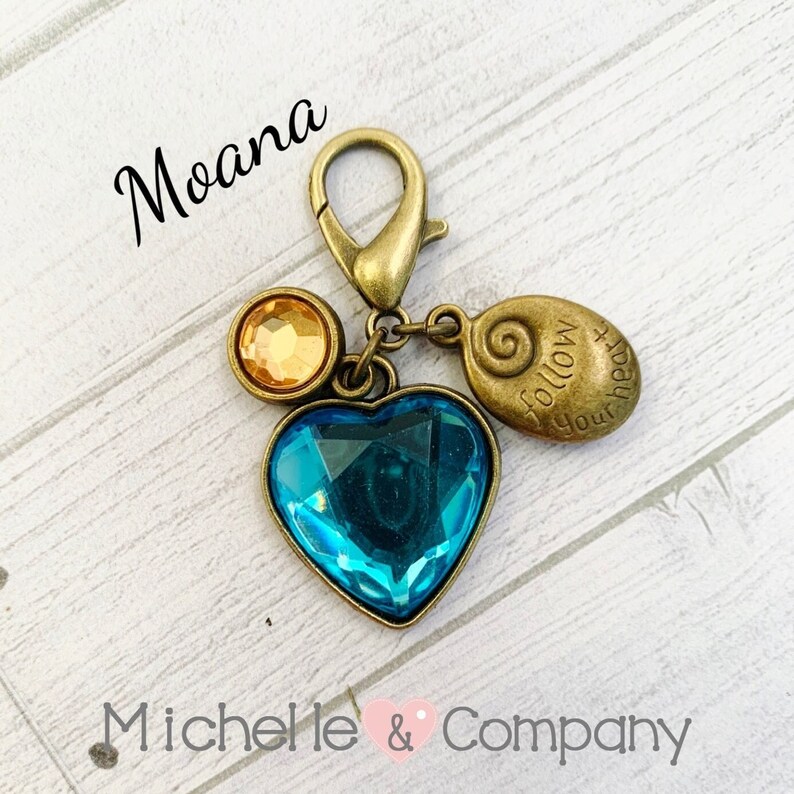 Op de afbeelding: Een gouden hanger met een blauw hart, twee gele strass steentjes en een hanger met de tekst "Volg je hart". De hanger is bevestigd aan een gouden ketting.