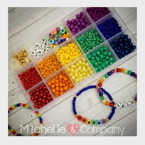 May include: A clear plastic container filled with colorful beads, including red, orange, yellow, green, blue, purple, and white. The beads are arranged in rows and columns, and there are also some gold beads and letter beads that spell out "HELLA" and "LELLA". Two bracelets are made with the beads, one with rainbow colored beads and the other with blue beads and gold stars.