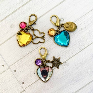 Op de afbeelding: Drie sleutelhangers met hartvormige kristallen. De eerste sleutelhanger heeft een geel hart, een roze edelsteen en een vlinderbedel. De tweede sleutelhanger heeft een blauw hart, een gouden edelsteen en een bedel met de tekst "Volg je hart". De derde sleutelhanger heeft een helder hart, een paarse edelsteen en een sneeuwvlokbedel.