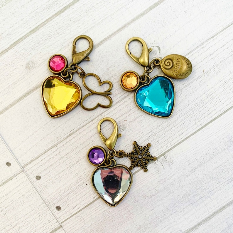 Op de afbeelding: Drie sleutelhangers met hartvormige kristallen. De eerste sleutelhanger heeft een geel hart, een roze edelsteen en een vlinderbedel. De tweede sleutelhanger heeft een blauw hart, een gouden edelsteen en een bedel met de tekst "Volg je hart". De derde sleutelhanger heeft een helder hart, een paarse edelsteen en een sneeuwvlokbedel.