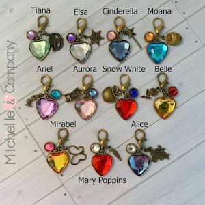 Op de afbeelding: Een set van 11 hartvormige sleutelhangers met verschillende gekleurde kristallen en bedels. Elke sleutelhanger heeft een andere Disney-prinses of een ander personage, waaronder Tiana, Elsa, Assepoester, Moana, Ariel, Aurora, Sneeuwwitje, Belle, Mirabel, Alice en Mary Poppins.
