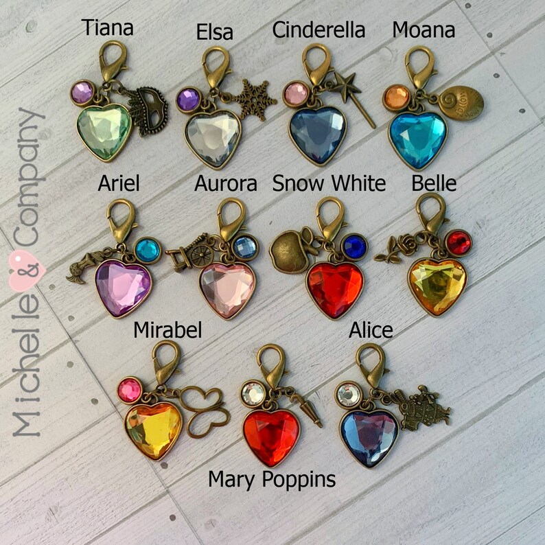 Op de afbeelding: Een set van 11 hartvormige sleutelhangers met verschillende gekleurde kristallen en bedels. Elke sleutelhanger heeft een andere Disney-prinses of een ander personage, waaronder Tiana, Elsa, Assepoester, Moana, Ariel, Aurora, Sneeuwwitje, Belle, Mirabel, Alice en Mary Poppins.