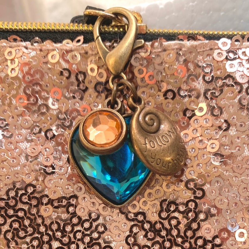 Op de afbeelding: Een gouden hanger met een blauw kristallen hart en een gouden hanger met de woorden "Volg je hart" aan een gouden ketting.