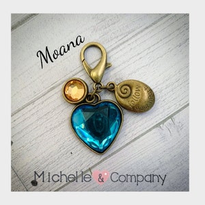 Op de afbeelding: Een gouden hanger met een blauw hartvormig kristal, een kleine gouden schelp met de woorden "Volg je hart" erop gegraveerd en een klein rond gouden kristal. De hanger is bevestigd aan een gouden karabijnhaak. De hanger is gelabeld met "Moana".