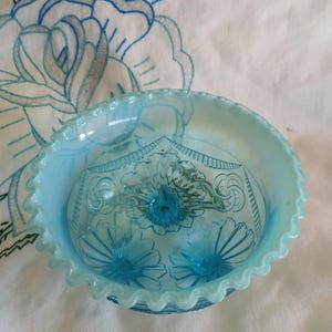 May include: A light blue, ruffled-edge glass bowl with a floral design. The bowl has a scalloped rim and a detailed pattern on the interior. The bowl is set against a white background with embroidered blue flowers.