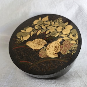 May include: A round, black decorative box with a detailed gold and cream-colored design. The design features two birds, flowers, and foliage. The box has a glossy finish and is likely made of lacquered wood.