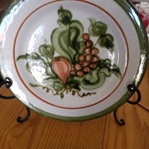 May include: A white ceramic plate with a green and brown floral design. The design features a cluster of grapes and leaves.