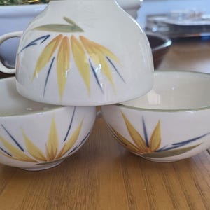 May include: Three white ceramic teacups with handles, stacked on a wooden surface. Each cup features a hand-painted design of a yellow and blue floral pattern. The cups are part of a tea set.