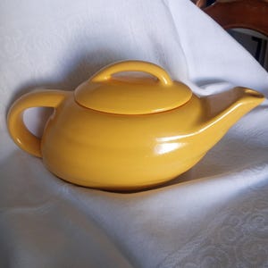 May include: A yellow ceramic teapot with a lid and a spout. The teapot is sitting on a white fabric surface.