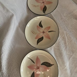 May include: Three off-white ceramic plates with a pink floral design and dark gray trim. Each plate features a pink flower with dark gray leaves and a dark gray rim. The plates are arranged in a vertical row.