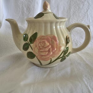 May include: Cream-colored ceramic teapot with a pink rose design and green leaves. The teapot has a curved spout, a rounded handle, and a lid with a small pink bud finial. The body of the teapot has vertical ridges.