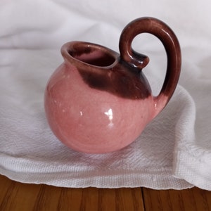 May include: A small, pink and brown ceramic pitcher with a rounded body and a curled handle.